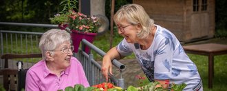 Zwei ältere Frauen unterhaltenEine Mitarbeiterin des Pflegedienstes unterhält sich mit einer älteren Frau. Beide lächeln während sie etwas außerhalb des Bildausschnitts betrachten. sich über einen Metallzaun hinweg. Im Vordergrund eine Pflanze die eine Schale Erdbeeren verdeckt.