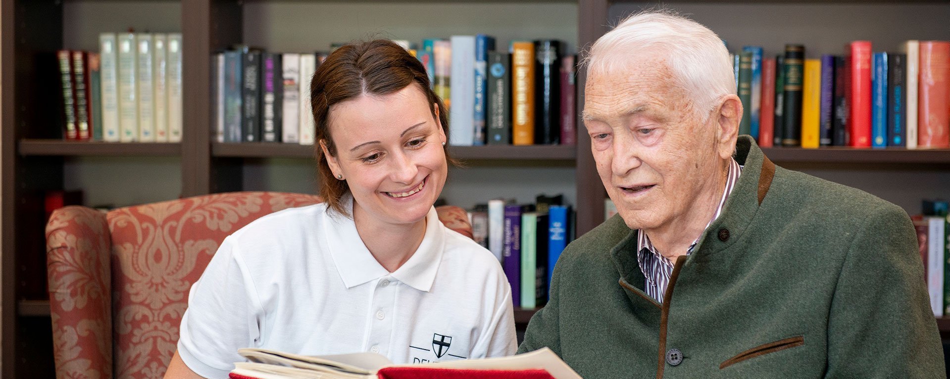 Ein älterer Herr zeigt einer Mitarbeiterin des Pflegedienstes eine Seite in einem rot eingebunden Buch. Im Hintergrund ein Bücherregal.