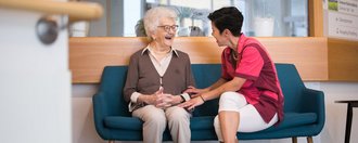 Eine Pflegerin und eine Bewohnerin sitzen auf einem Sofa, unterhalten sich und lachen