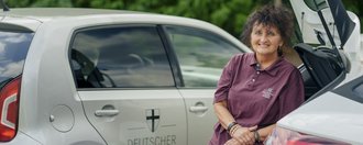 Eine Mitarbeiterin des ambulanten Pflegedienstes lehnt an ihrem Dienstfahrzeug und lächtelt in die Kamera.