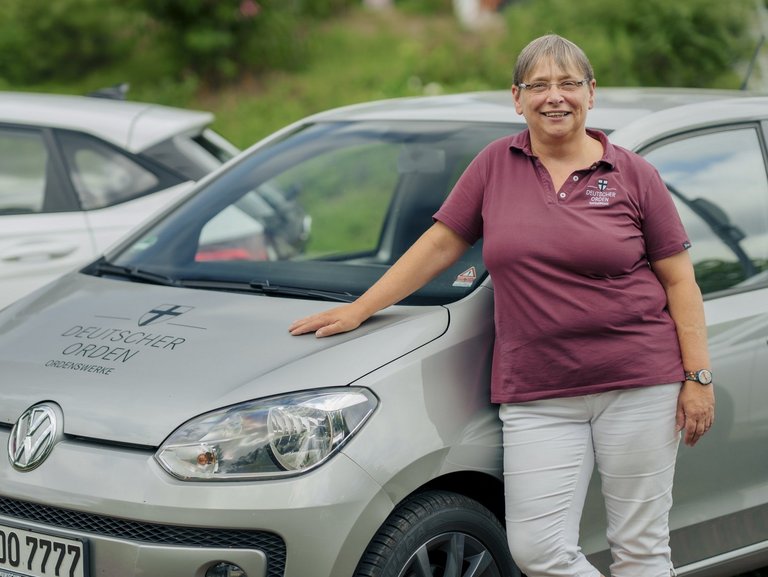 Eine Mitarbeiterin des ambulanten Pflegedienstes lehnt an ihrem Dienstfahrzeug und lächtelt in die Kamera.